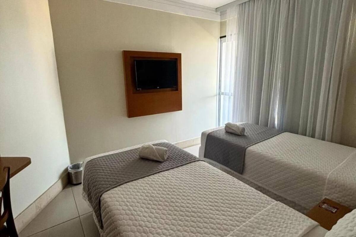 A hotel room with two beds, a TV mounted on the wall, and a window with curtains.