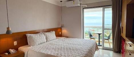 A hotel room with a large bed, a nightstand, an air conditioning unit, a TV, and a balcony with a view.