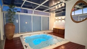 A hot tub in a covered outdoor area with a wooden deck, a large potted plant, and a wall-mounted mirror. - Royal Urban Macaé Hotel (Macaé)