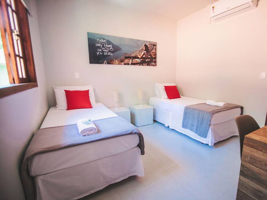 A hotel room with two single beds, each with a red pillow and a rolled towel. There is a small table and a chair, a wall-mounted air conditioner, and a painting above the beds.