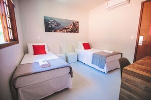 Habitación de hotel con dos camas individuales, cada una con un almohadón rojo y una toalla enrollada. Hay una mesita y una silla, un aire acondicionado de pared y un cuadro sobre las camas.