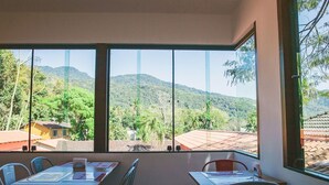 Una habitación con amplios ventanales que ofrecen vistas a un paisaje montañoso y un pueblo. Hay dos mesas con sillas, una con un menú y la otra con un cuaderno.