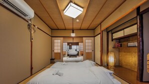 A traditional Japanese-style room with tatami flooring, a futon bed, a shoji screen, and a wooden ceiling. - Shiki Homes SHINJIN (Kyoto)