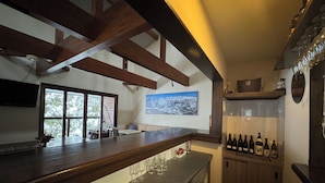 A bar area with a wooden counter, a refrigerator stocked with bottles, and a large window with a view of snow-covered trees. - Bears Den Mountain Lodge (Hakuba)