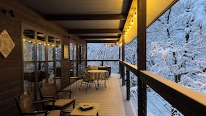A wooden deck with outdoor seating, a glass wall, and a view of snow-covered trees. - Bears Den Mountain Lodge (Hakuba)