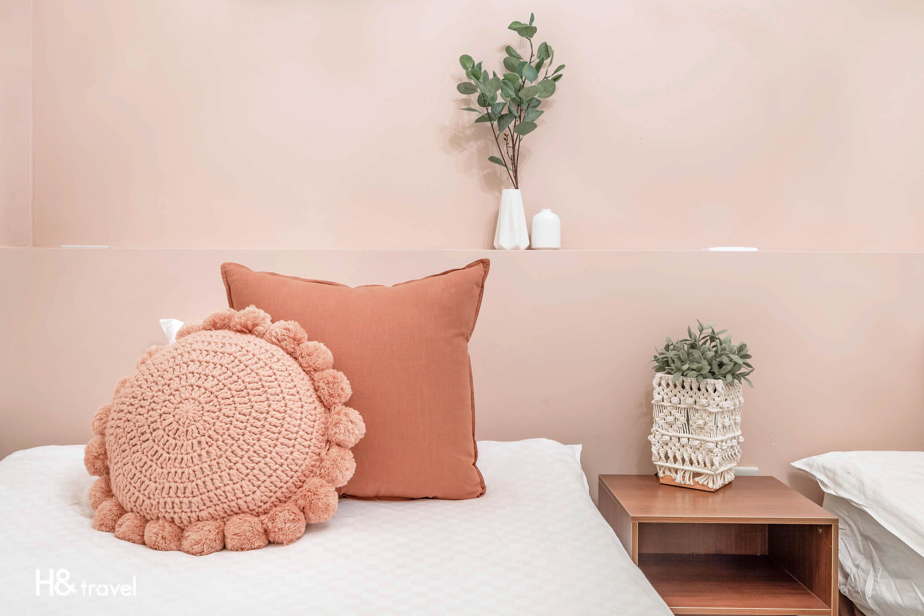 Un dormitorio ordenado con un cojín grande y redondo de ganchillo, un cojín de color liso y una mesita de noche con una planta.