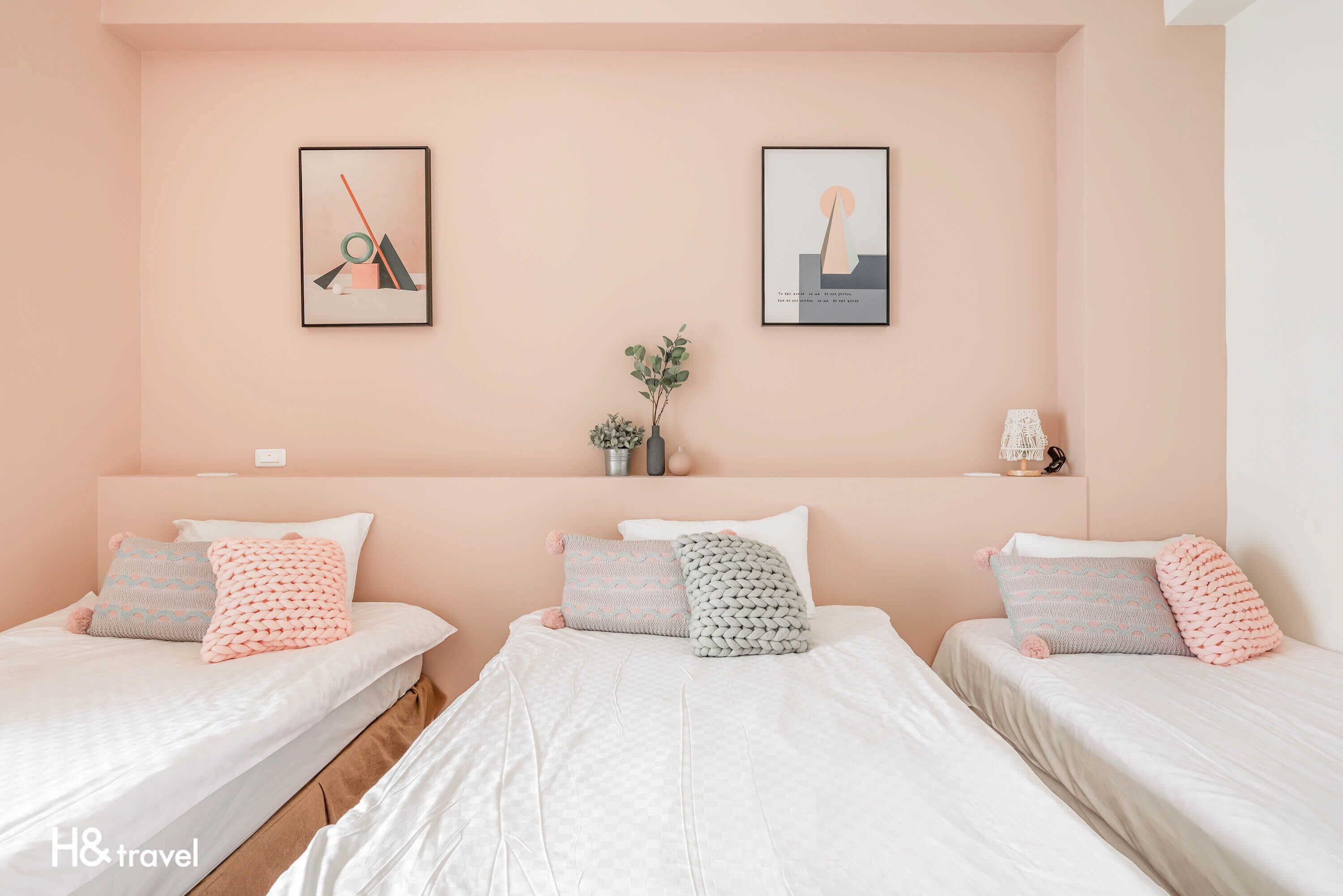 A hotel room with three beds, each with a white mattress and pink and grey patterned pillows. There are two framed artworks on the wall, a small plant on the nightstand, and a white lamp.