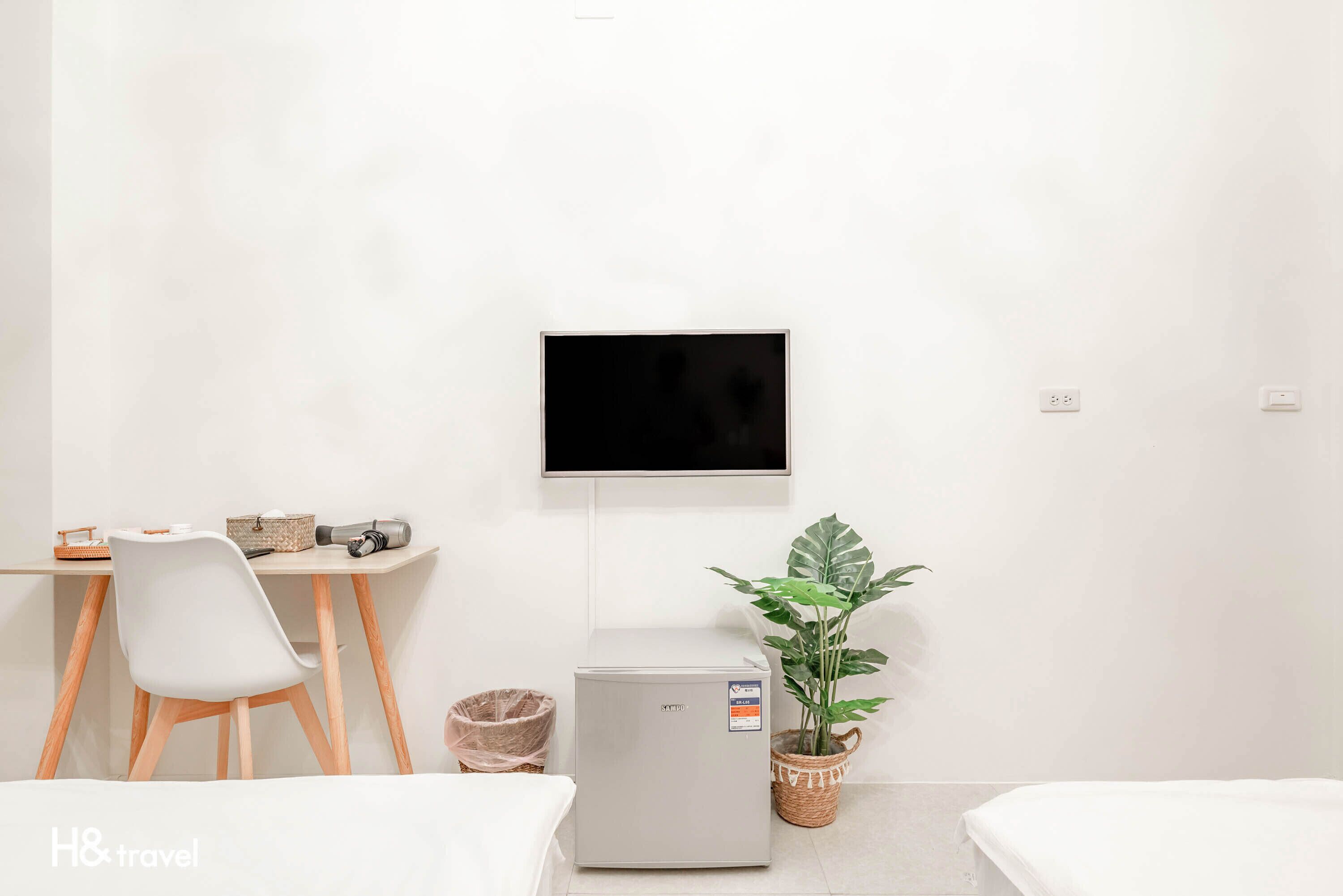 Quarto de hotel com uma cama, uma escrivaninha com cadeira, um frigorífico pequeno, um vaso de plantas e uma televisão na parede.