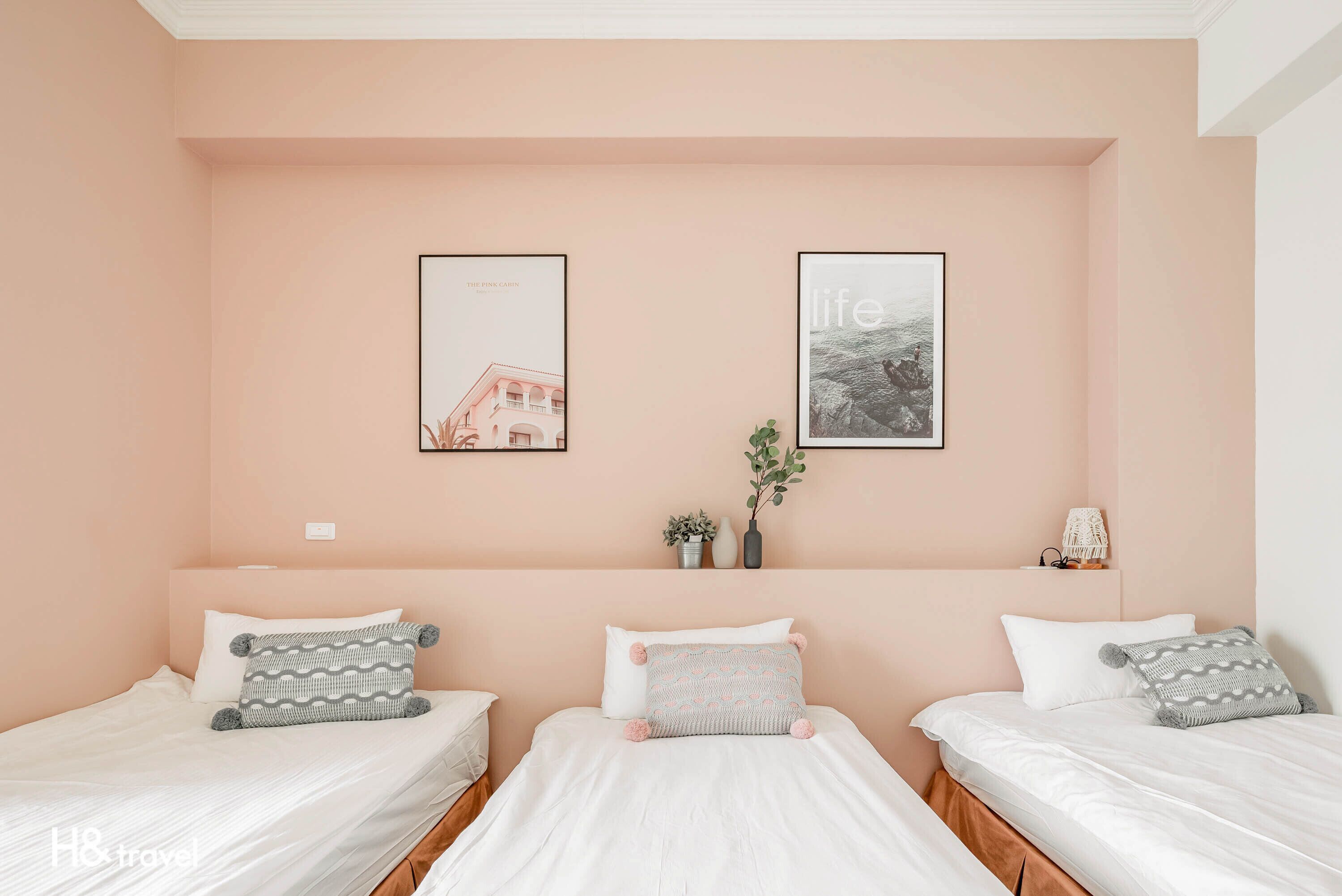A hotel room with three beds, each with white bedding and decorative pillows. There are two framed pictures on the wall, one depicting a building and the other a landscape.