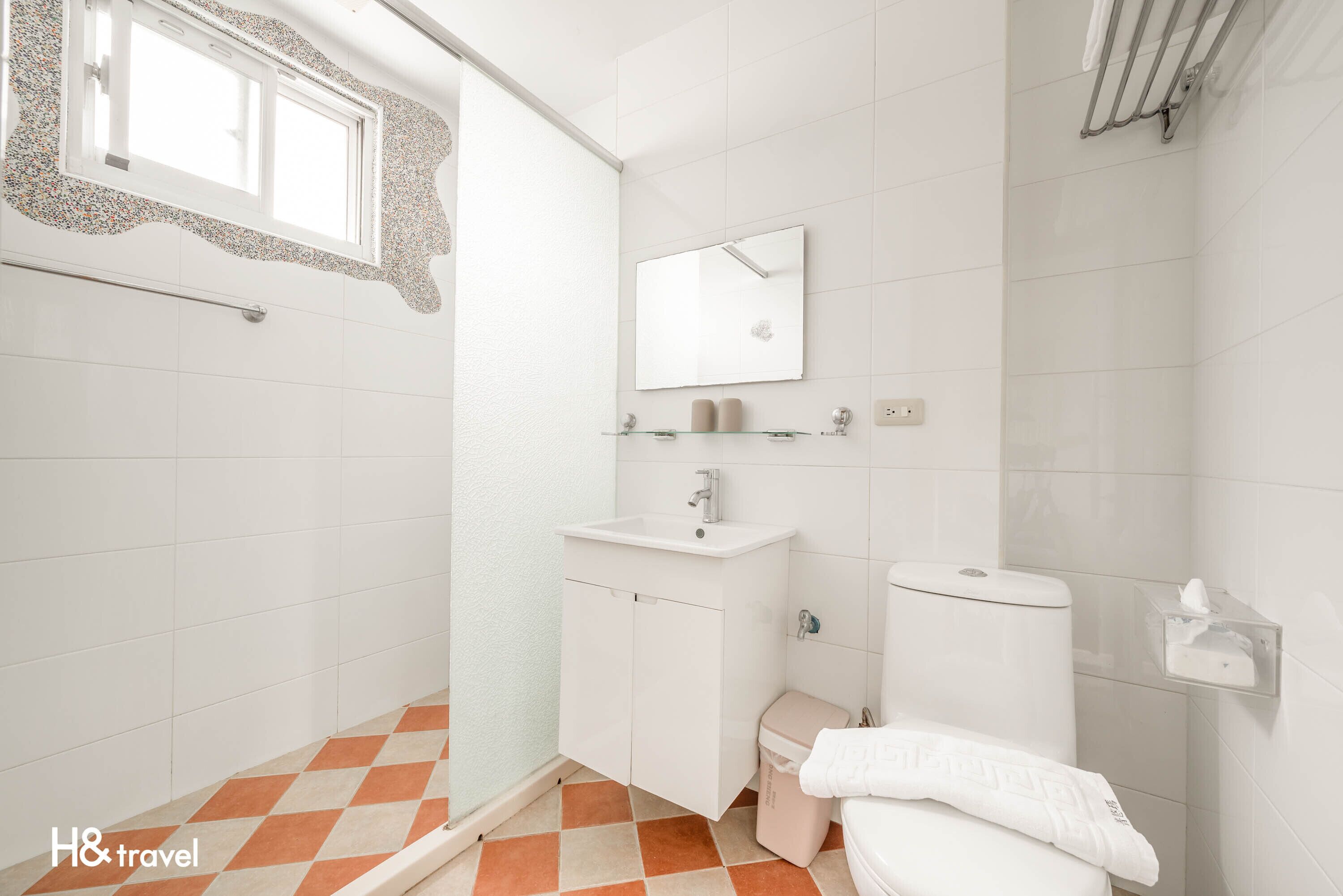 Une salle de bain avec un lavabo blanc, un miroir et des toilettes. Il y a une fenêtre avec un cadre décoratif, et le sol est recouvert d’un motif à carreaux.