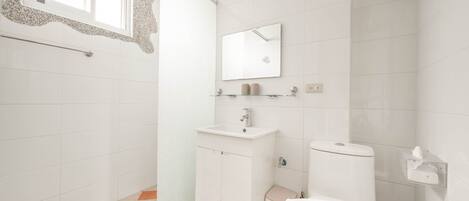 Une salle de bain avec un lavabo blanc, un miroir et des toilettes. Il y a une fenêtre avec un cadre décoratif, et le sol est recouvert d’un motif à carreaux.