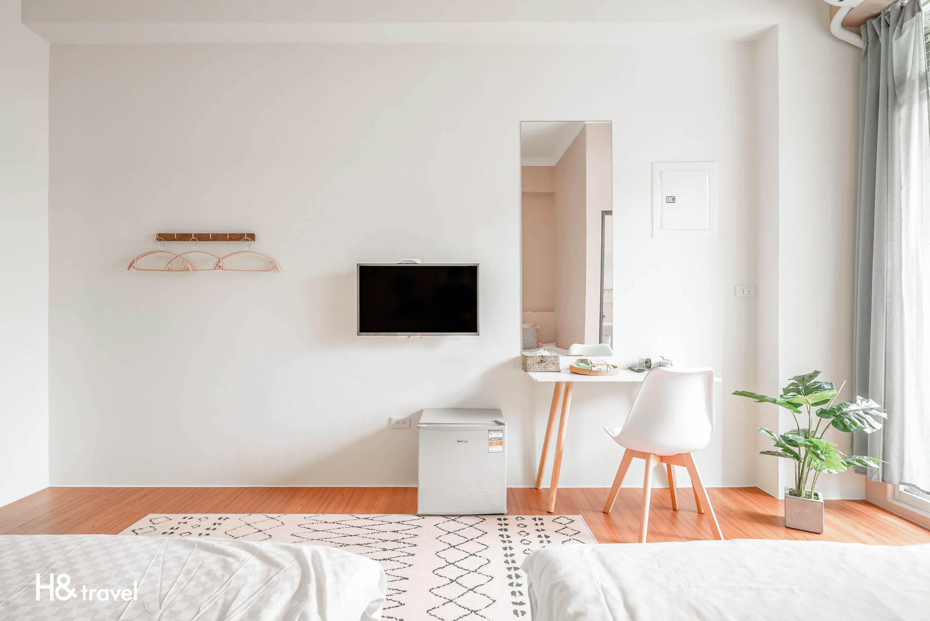 A modern hotel room with a bed, a desk, a television, a small refrigerator, and a potted plant.