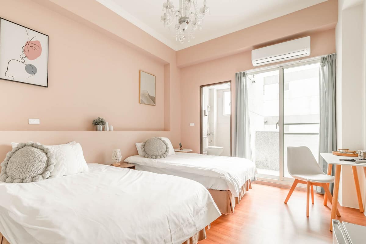 Une chambre d’hôtel avec deux lits, un bureau avec une chaise, un lustre et une grande fenêtre avec des rideaux.