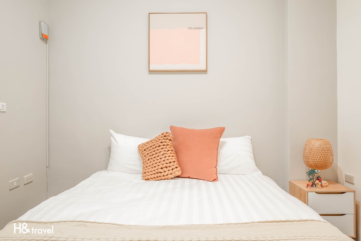 Une chambre d’hôtel bien rangée, avec un lit, des oreillers, une lampe de chevet et un tableau encadré au mur.