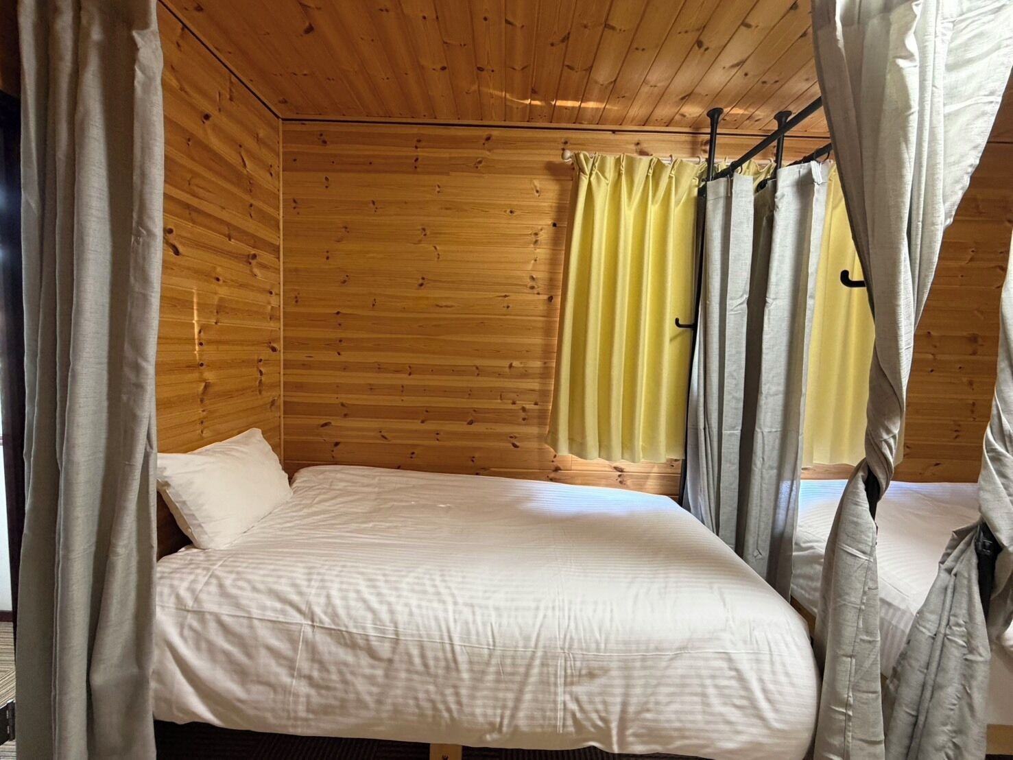 A wooden cabin room with a bed, curtains, and a window with yellow curtains.
