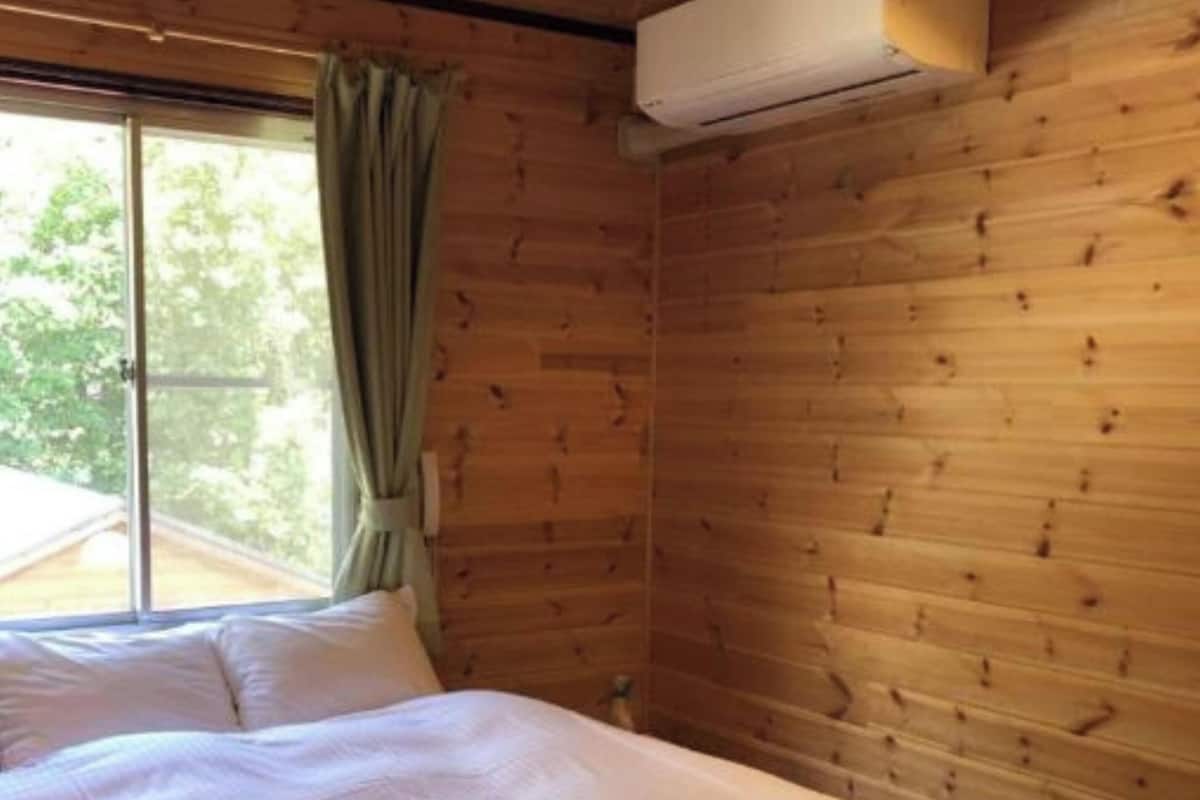 Ein Schlafzimmer mit Bett, einem Fenster mit Vorhängen und einer Klimaanlage. Es wird angegeben, dass das Badezimmer mit anderen geteilt wird.