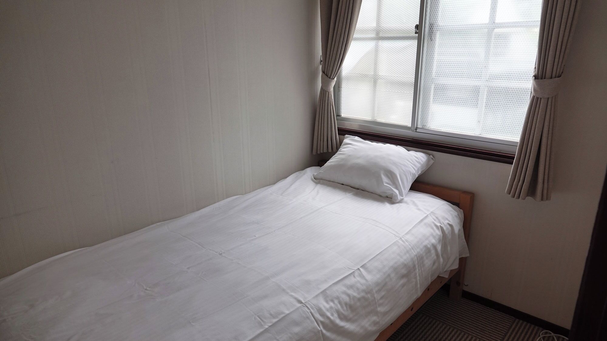 A single bed with white bedding in a room with a window and curtains.