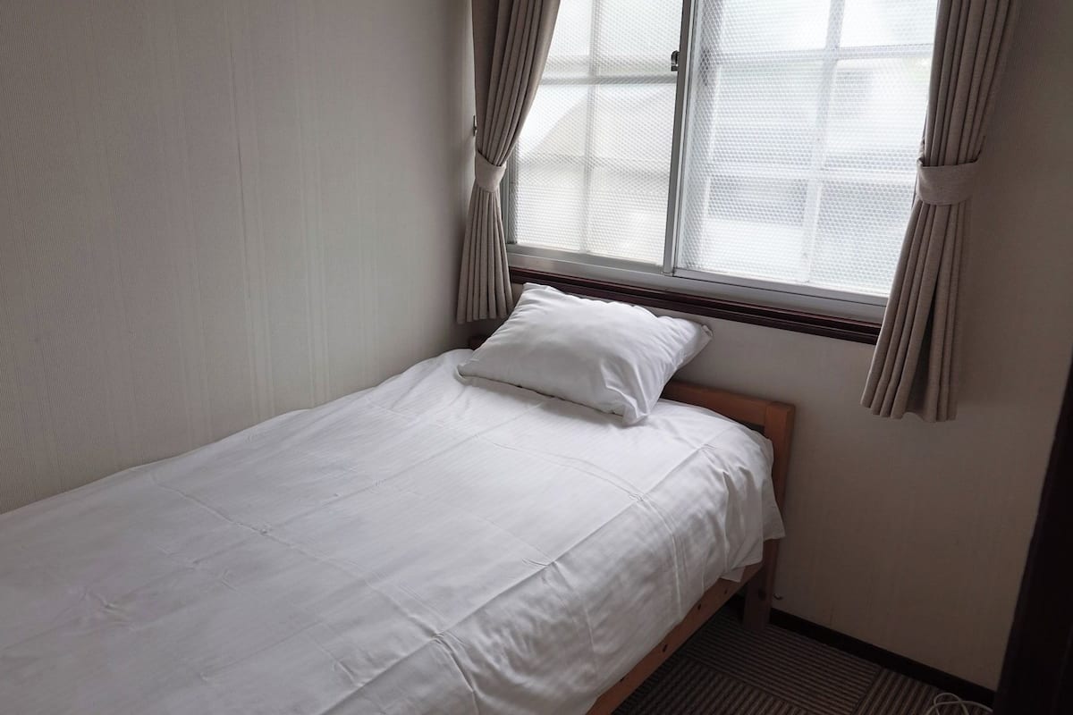 Ein Einzelbett mit weißer Bettwäsche in einem Zimmer mit Fenster und Vorhängen.