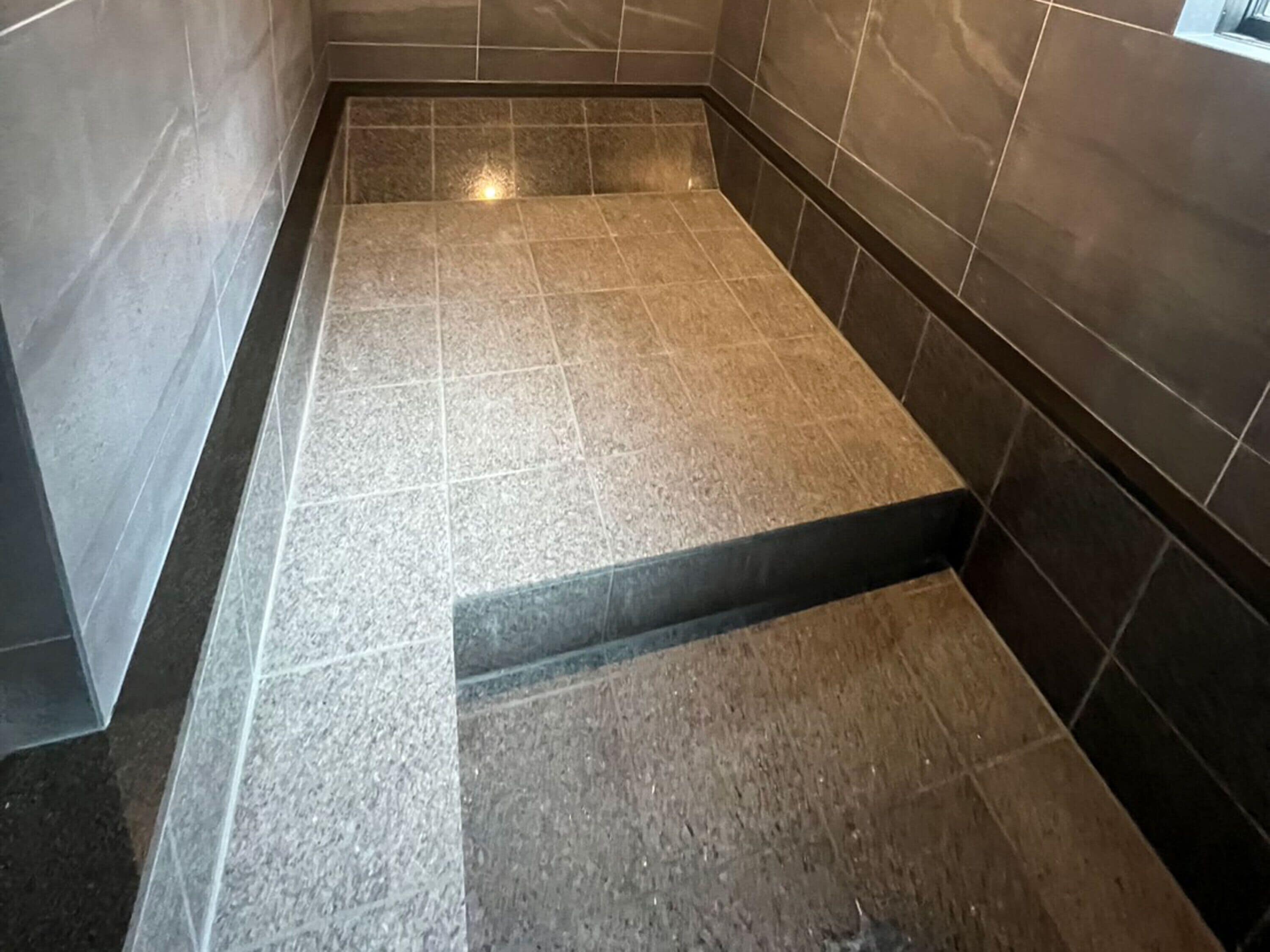 Zona de ducha con azulejos, suelo y paredes marrón oscuro, desagüe visible e iluminación sobre ella.
