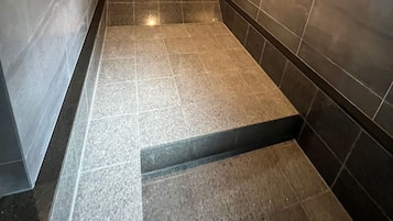Zona de ducha con azulejos, suelo y paredes marrón oscuro, desagüe visible e iluminación sobre ella.