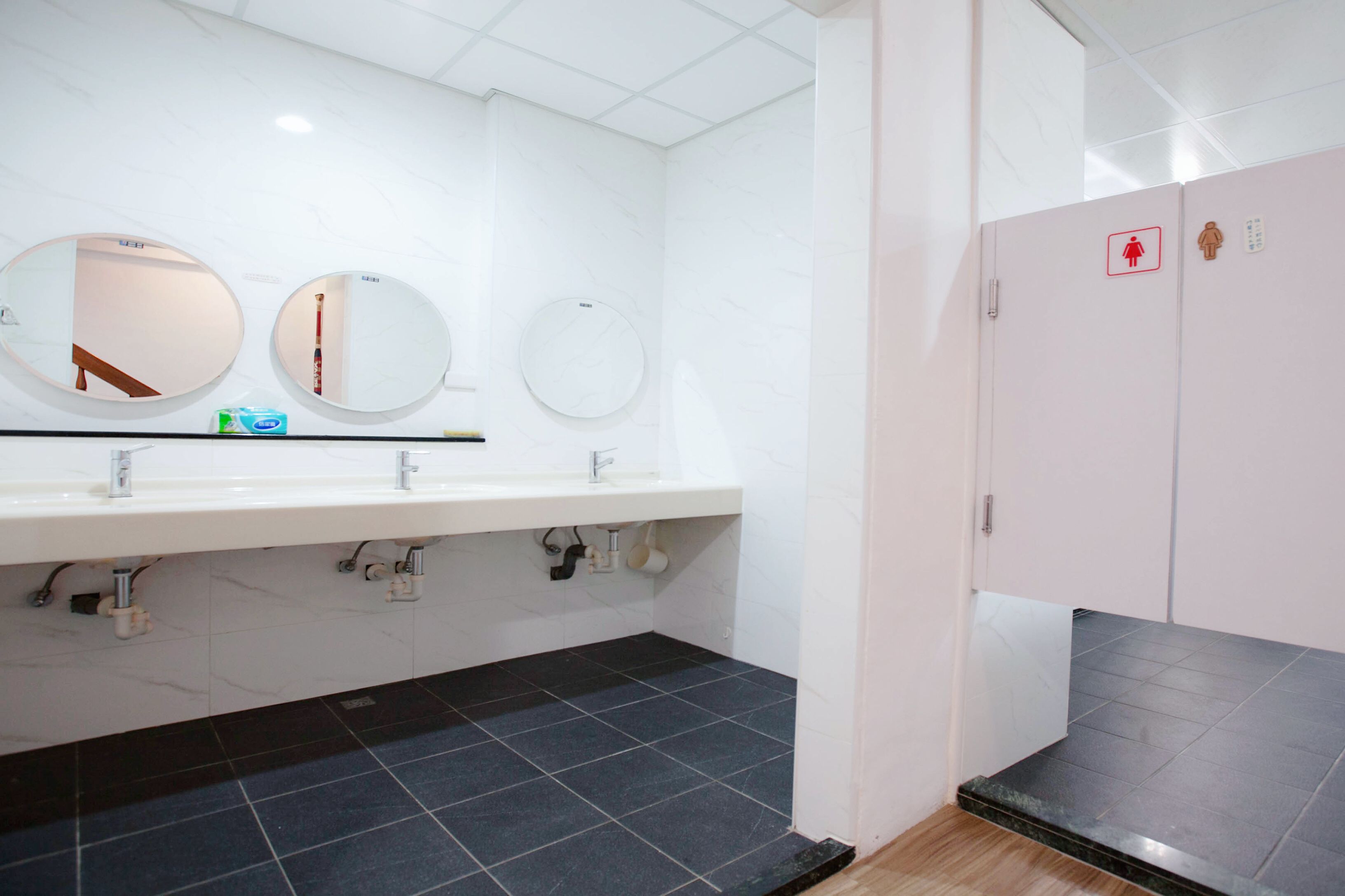 A public restroom with three round mirrors, a white sink with a soap dispenser, and a black tiled floor.