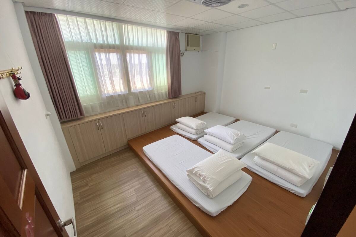 A hotel room with three single beds, a wooden floor, a window with curtains, a radiator, and a door with a handle.