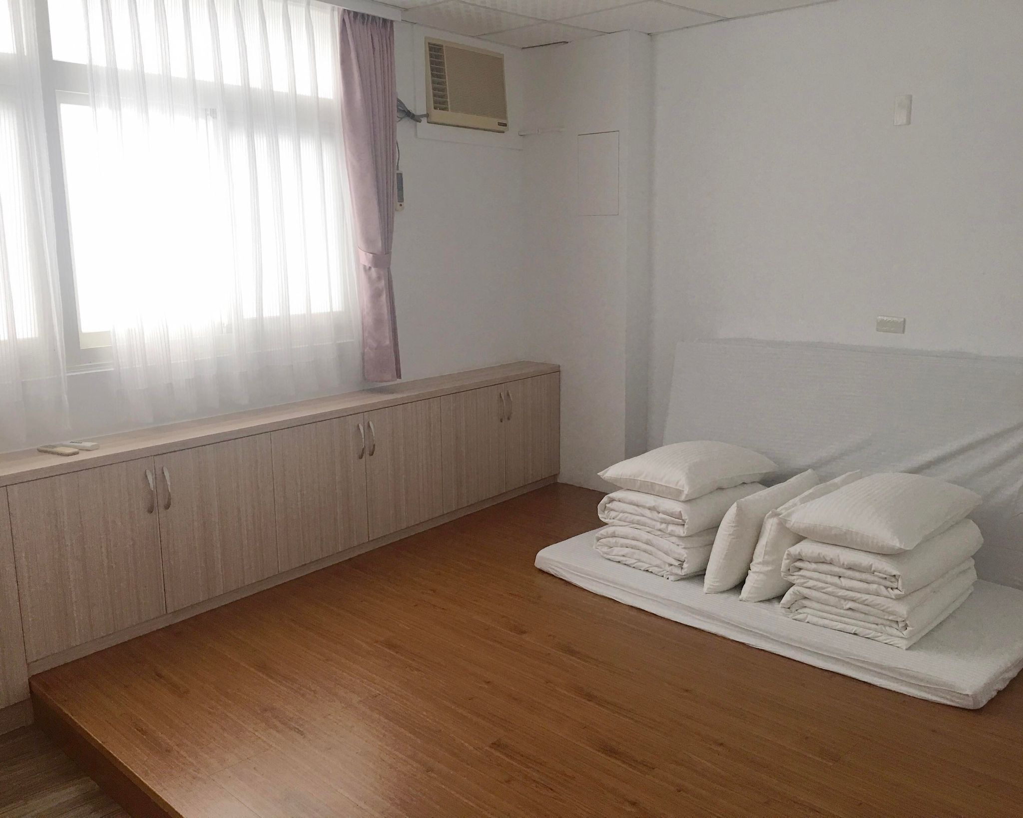 A room with a wooden floor, a window with sheer curtains, a radiator, and a bed with neatly folded white linens.