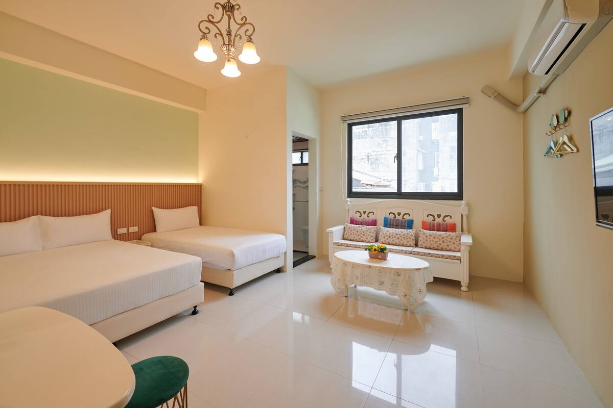 A modern hotel room with two beds, a sofa, a small table, and a large window.