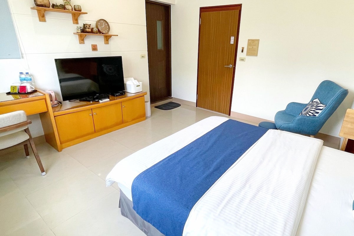 A hotel room with a bed, a desk with a TV, a chair, and shelves with decorative items.