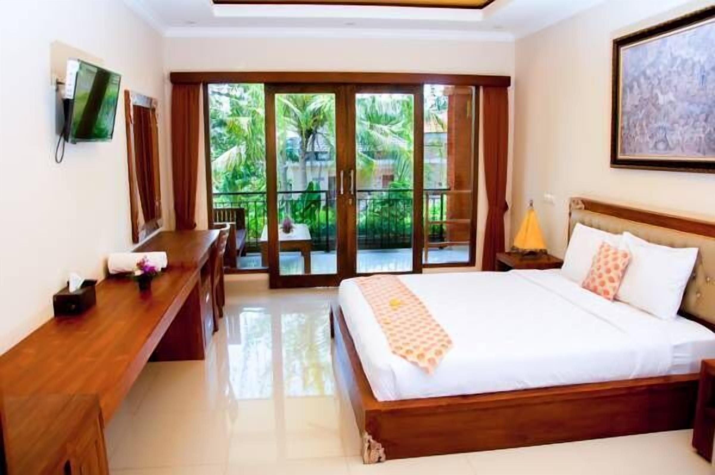 A hotel room with a bed, a wooden desk, a TV, and a balcony with a view of greenery.