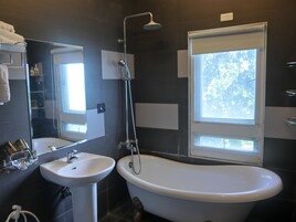 Un baño moderno con una bañera independiente, un lavabo sobre pedestal y un espejo grande. Hay una ventana que proporciona luz natural y vistas a zonas verdes.