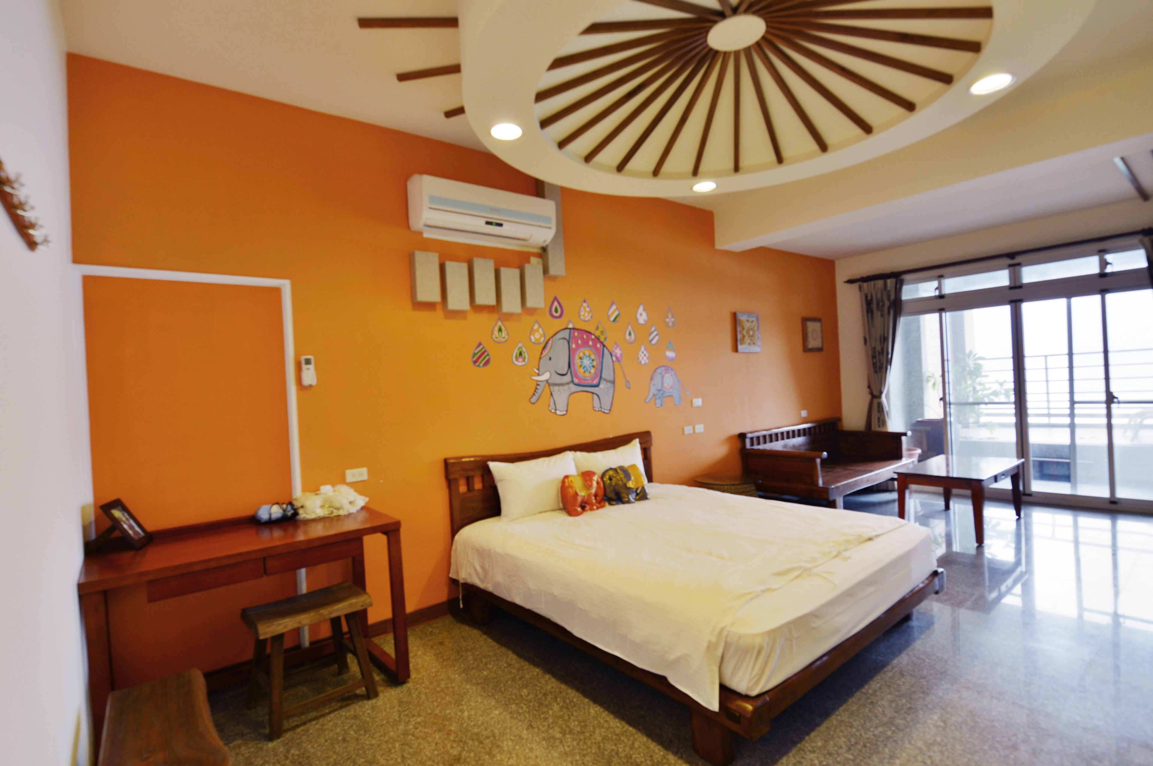 A hotel room with a large bed, a desk, and a chair. There is an air conditioning unit on the wall and a window with curtains.