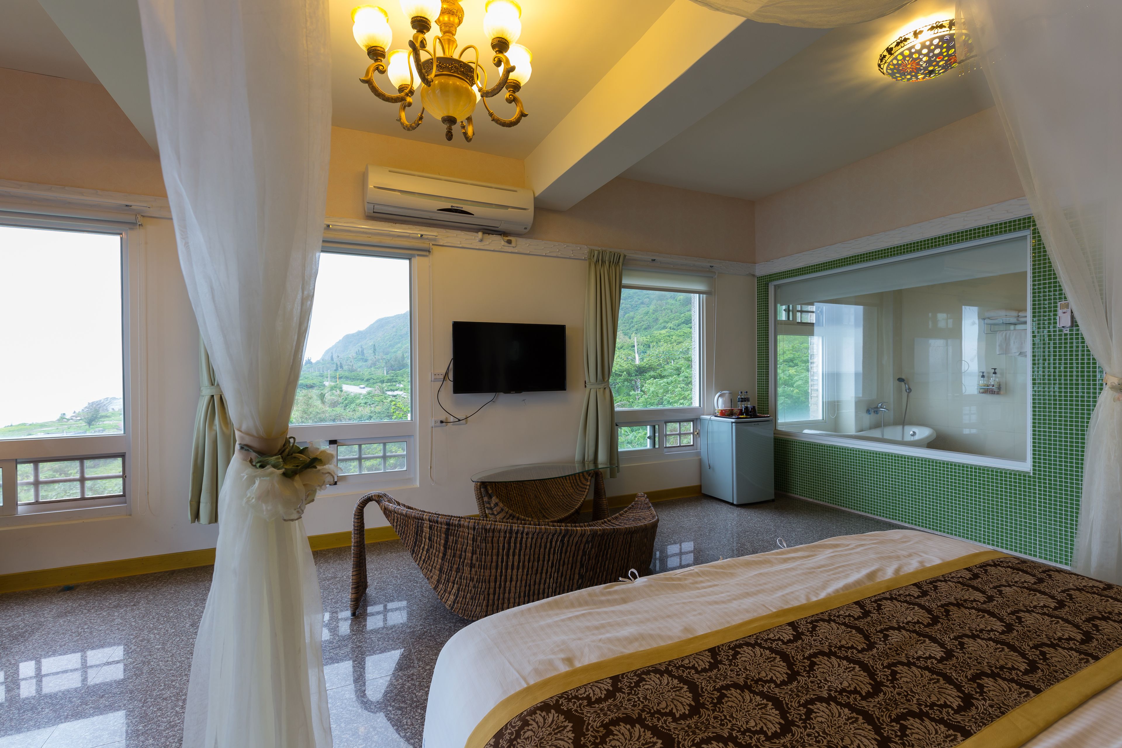 A hotel room with a bed, a television, a window with a view of greenery, and a bathroom with a green-tiled wall.