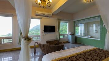 A hotel room with a bed, a television, a window with a view of greenery, and a bathroom with a green-tiled wall.
