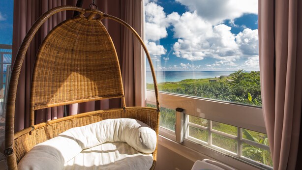 Una silla de mimbre con cojín junto a una ventana con vistas al océano y vegetación.