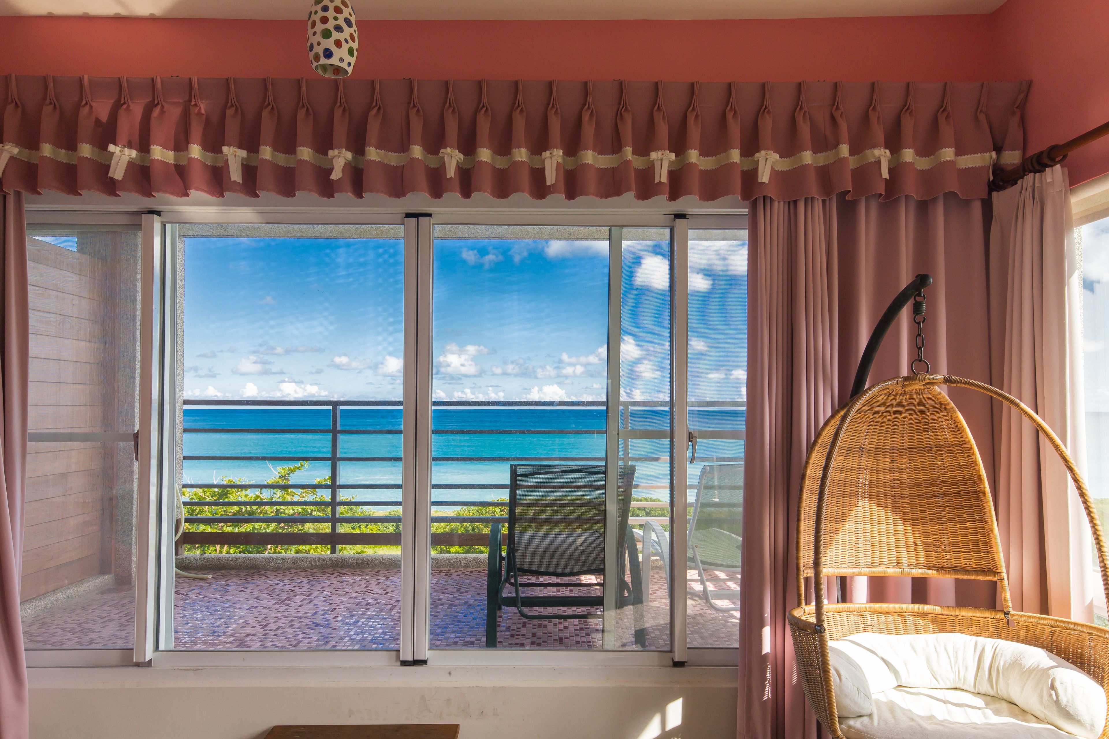 A room with a large sliding glass door leading to a balcony with a view of the ocean. There is a wicker chair with a cushion on the balcony.