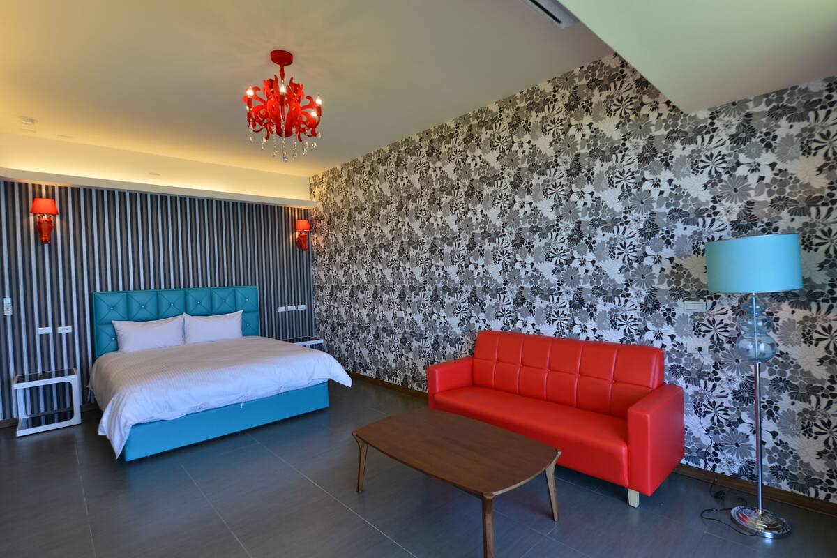 Un dormitorio con una cama, un sofá rojo, una mesa de centro de madera, una araña y un papel tapiz estampado.