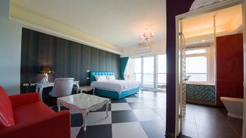 A hotel room with a checkered floor, a bed with blue bedding, a red armchair, a white table, and a bathroom visible through an open door.