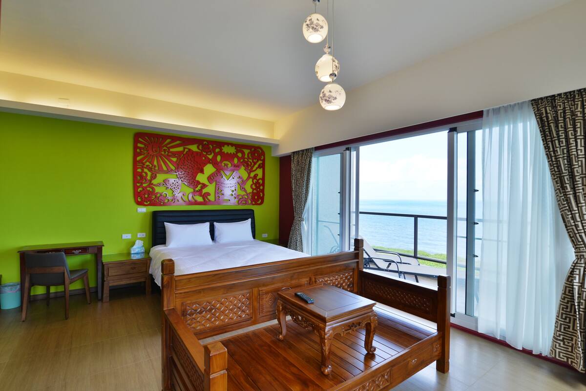 Habitación de hotel con una cama grande, un escritorio de madera, una silla, una mesita y vistas al mar a través de amplios ventanales.