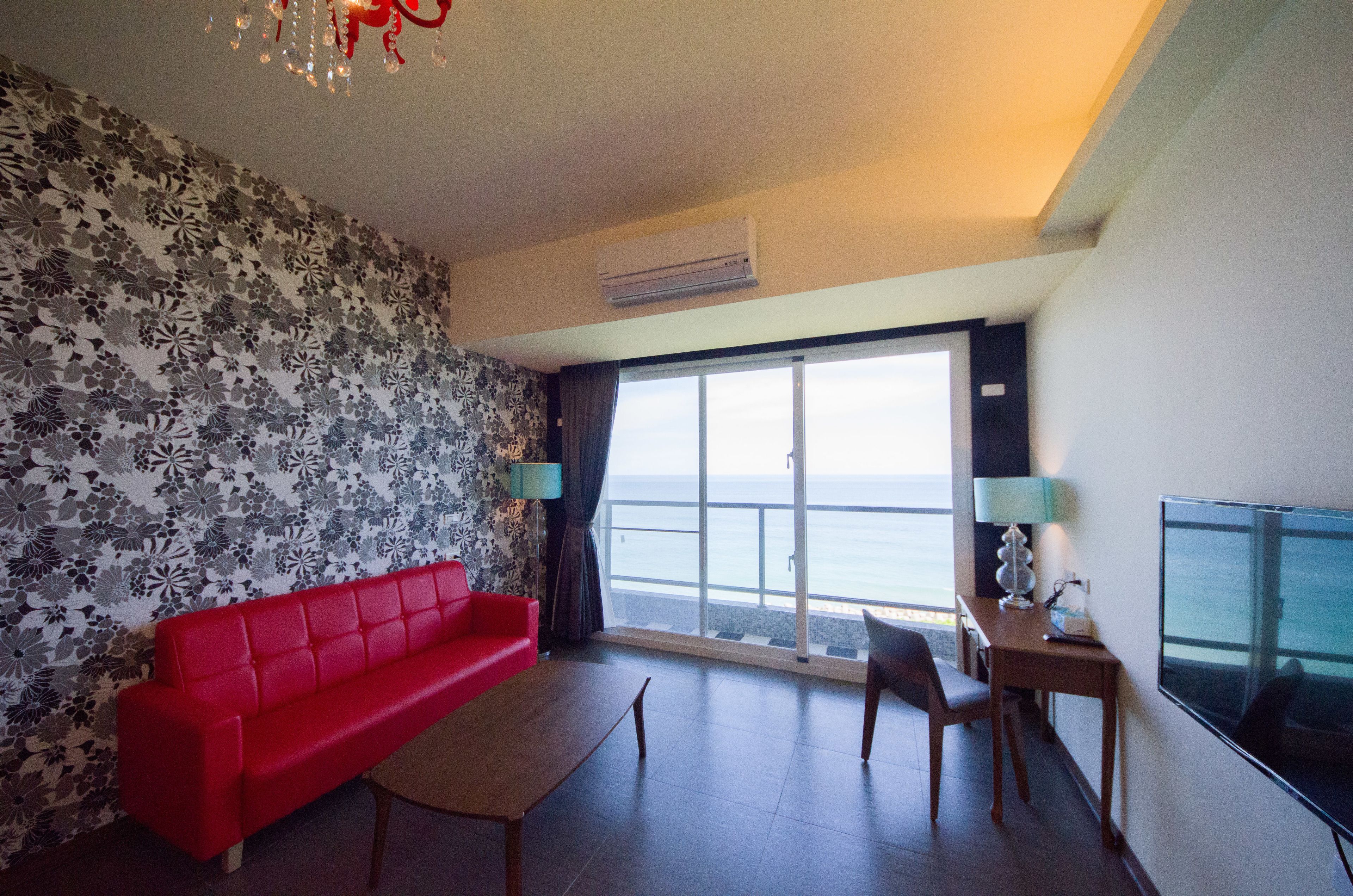 Quarto com sofá vermelho, uma mesa pequena, televisão e uma janela grande com vista para o mar.