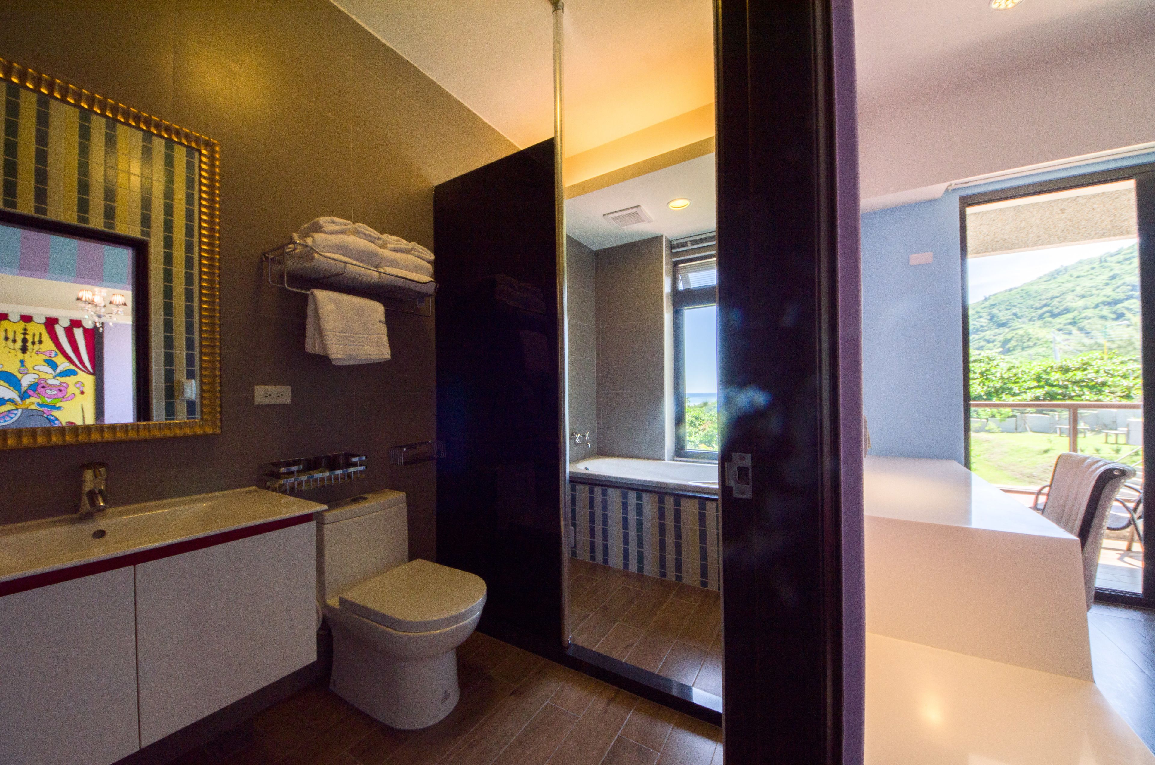 A modern bathroom with a toilet, sink, and towel rack. There is a window leading to a balcony with a view of greenery.