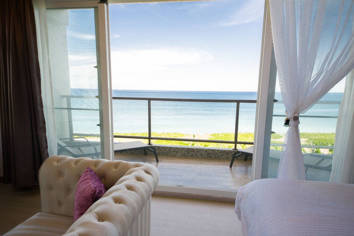 Un dormitorio con un amplio ventanal con vistas al mar, una cama, un sillón con cojines y un balcón con barandilla de cristal.