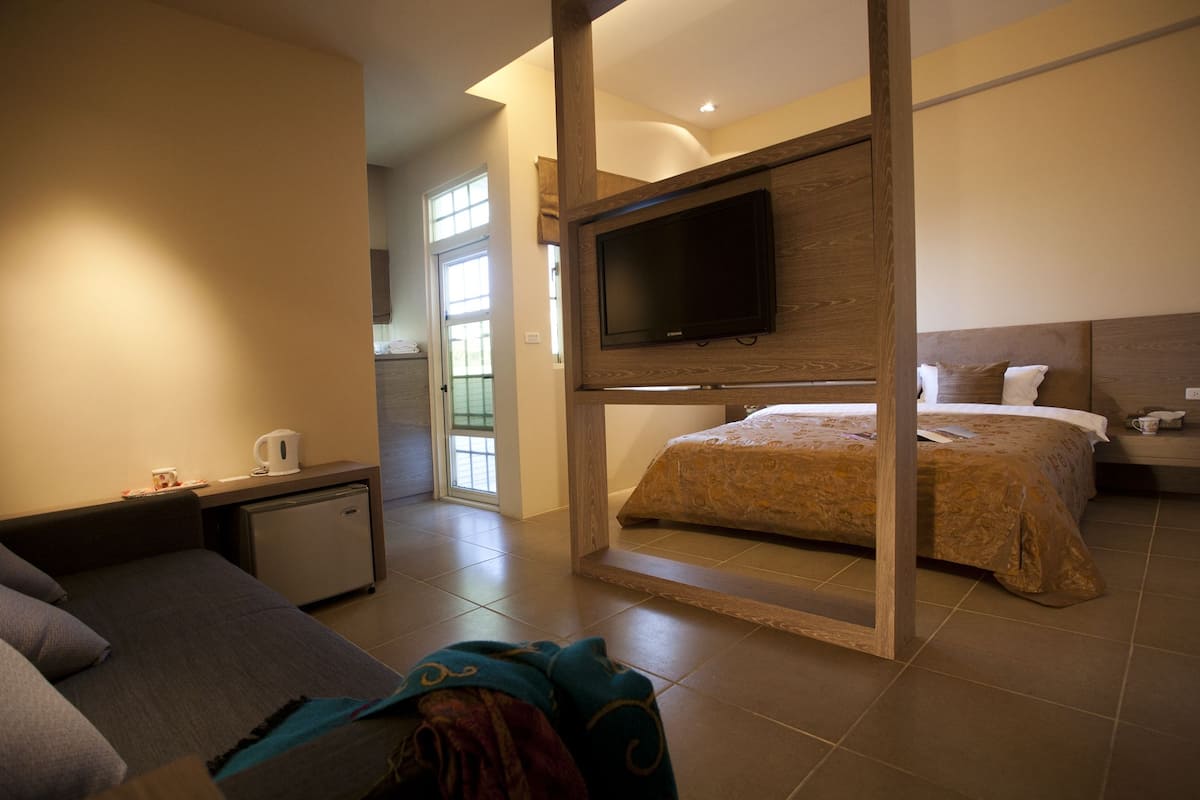 A hotel room with a bed, a TV mounted on the wall, a desk with a chair, and a small kitchenette area.