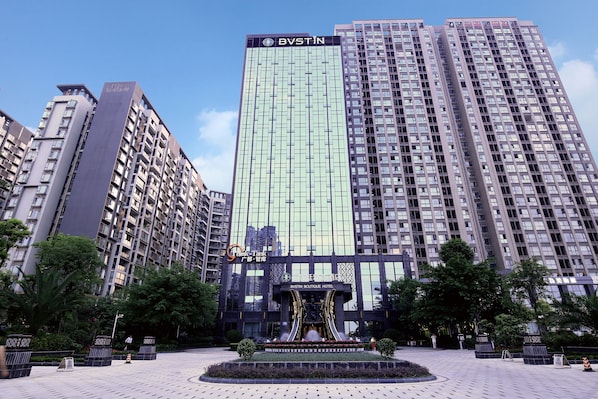 A modern high-rise building with a reflective glass facade, a central entrance with decorative elements, and a landscaped plaza with a fountain.