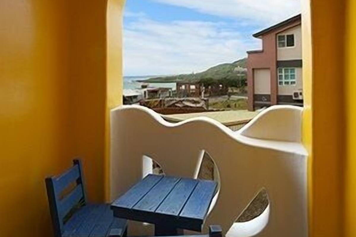 A room with a yellow wall, a window with a view of a beach and buildings, a table, and two chairs.