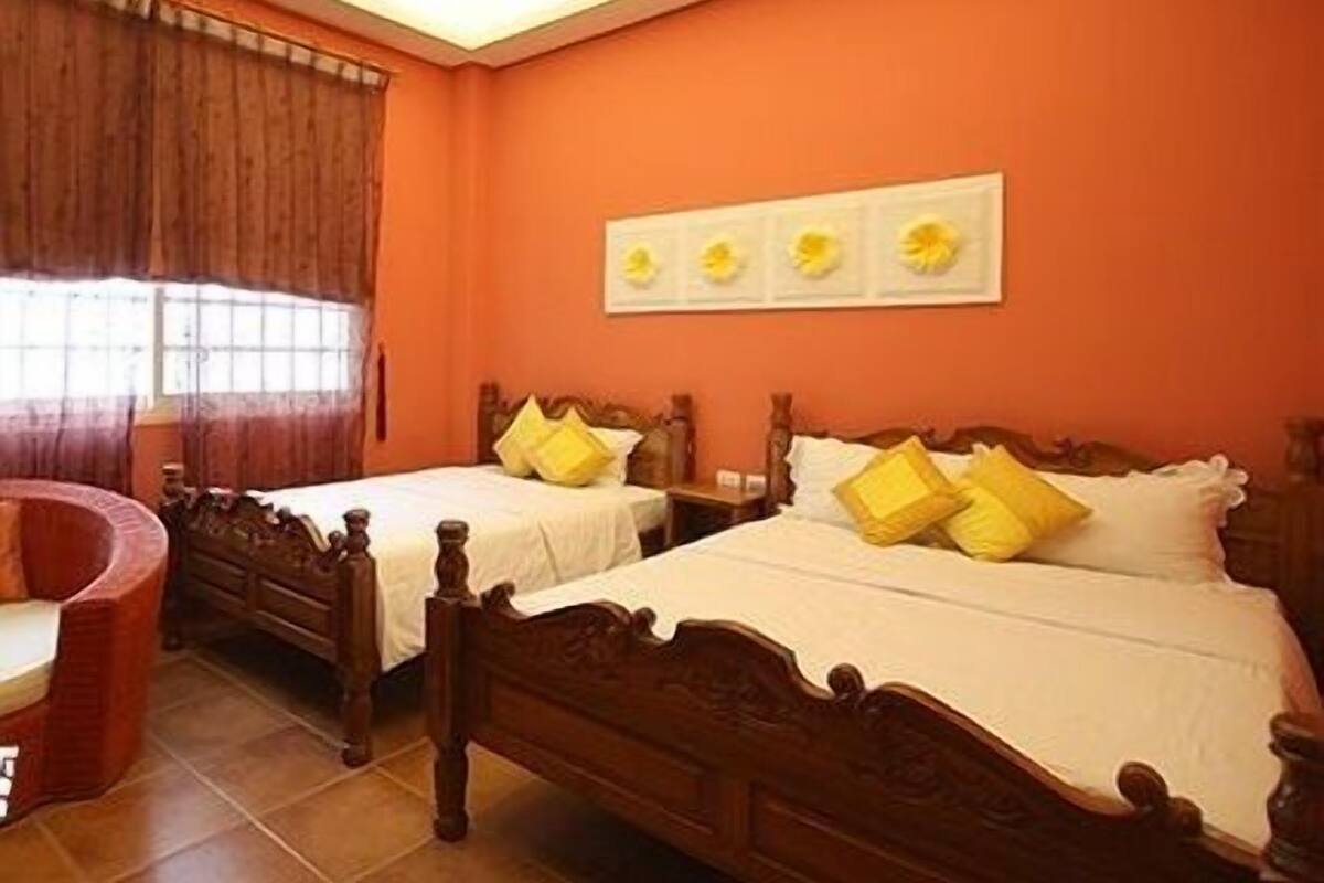 A room with two wooden beds, each with white bedding and yellow pillows. There is a window with curtains, a wall-mounted artwork, and a red chair.
