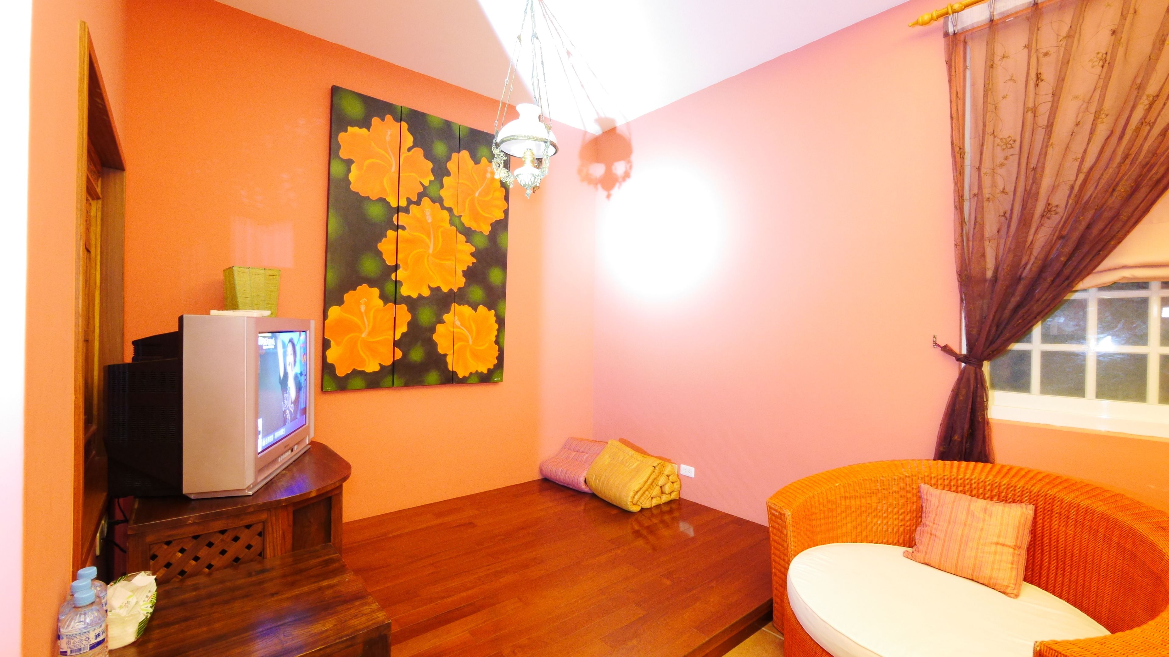 Una habitación con piso de madera, un televisor sobre un mueble de madera, un cuadro de flores amarillas, un sofá de mimbre con un cojín y una ventana con cortinas.