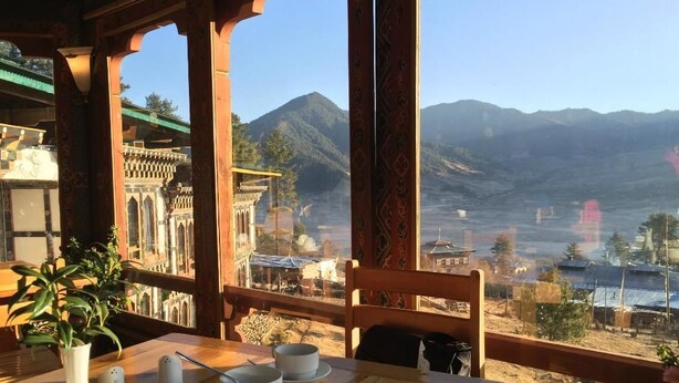 Una mesa de comedor con vajilla y tazas, un florero con una planta, y vistas a montañas y un pueblo.