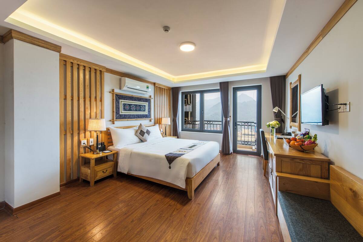 Ein Hotelzimmer mit einem Holzbett, Nachttischen, einem Schreibtisch mit Lampe, einem Fernseher und einem Balkon mit Aussicht.