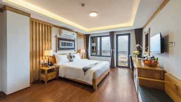 A hotel room with a wooden bed, bedside tables, a desk with a lamp, a TV, and a balcony with a view.